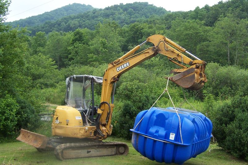 Excavator lifting a blue polyethylene septic tank into place